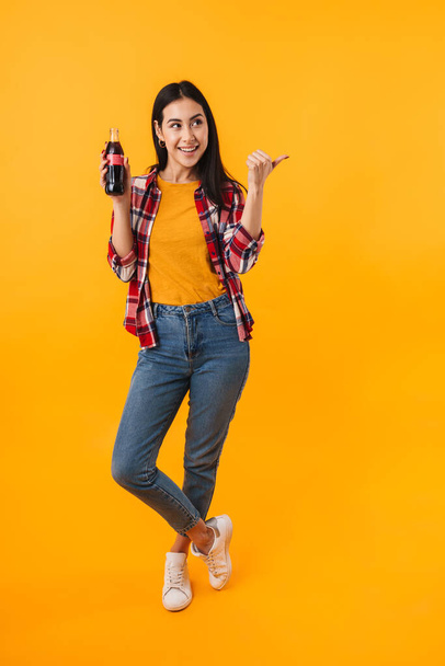Photo of cheerful nice woman pointing finger aside and drinking soda isolated over yellow background - Zdjęcie, obraz