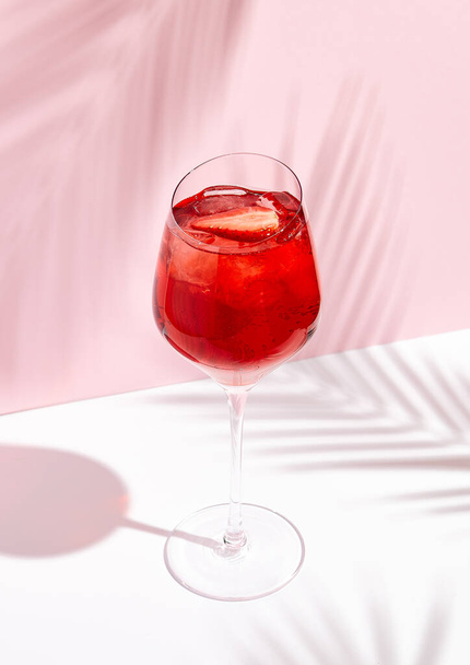Red summer cocktail with strawberry in wine glass on creative background. Shadows of palm leaf on pink wall and white floor.  - Фото, зображення