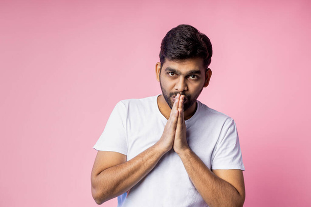 Young religious indian unshaven man wearing white t shirt, keeping palms together, looking down, praying, hoping for best, isolated over in pink background. Belief concept. - Фото, изображение