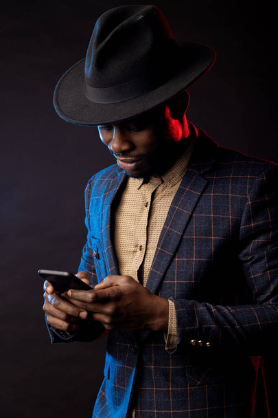 Portrait of confident stylish young african man in formalwear, holding mobile phone, looking at screen, standing against black background. Successful businessman. Business, technology. - Zdjęcie, obraz