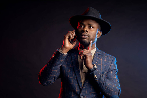 Horizontal shot of serious worried stylish young african man wearing suit, hat, talking on cell phone, standing in dark against black background . Successful businessman. - Foto, Imagem