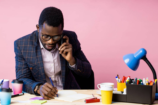 Serious afro american businessman in formal wear, glasses, talking on mobile phone, looking at notepad, holding pen, sitting at desk in office. Job, business concept. - Foto, Bild