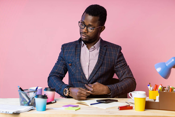 Horizontal shot of handsome confident businesslike afro american businessman sitting at workplace in office, fastening button on suit, wearing eyewear, posing over pink wall. - Foto, Bild