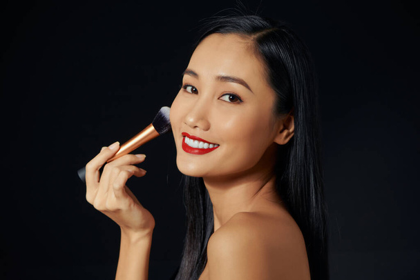 Schöne junge Asiatin benutzt weichen Pinsel und Kosmetik auf ihrem eigenen Gesicht.  - Foto, Bild