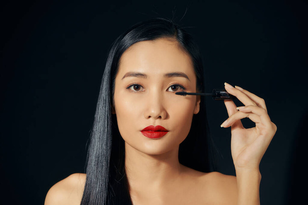 Primer plano retrato de bonito lindo dulce atractivo alegre positivo asiático mujer sosteniendo en las manos aplicando rímel negro de moda aislado
 - Foto, imagen