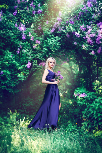 Outdoor fashion photo of a beautiful young woman surrounded by purple flowers. Girls in a long dress with a slit on the background of a spring garden with lilacs. The concept of cosmetics and perfumes. Lilac bouquet and flowers in your hair - Fotografie, Obrázek
