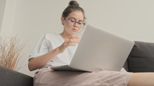 Vista desde abajo de la bonita mujer morena concentrada en gafas usando computadora portátil mientras está sentada en el sofá en casa
 - Metraje, vídeo