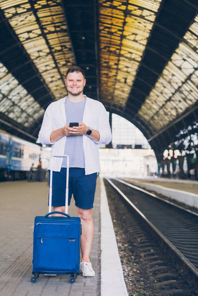 homme debout avec valise à la gare attendant le train en utilisant un téléphone portable surf internet
 - Photo, image