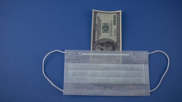 Tıbbi bir maskenin içinde mavi bir arka planda 100 dolarlık banknot. - Fotoğraf, Görsel