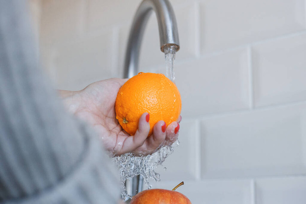 Κοντινό πλάνο της νεαρής γυναίκας πλένει τα φρούτα για να αποτρέψει τη μόλυνση από τον ιό της στέψης. Το πορτοκάλι και το μήλο πλένονται αγνά για να διατηρηθεί η υγιεινή. Ασφάλεια ιών. Έννοια διακοπής Covid-19. - Φωτογραφία, εικόνα