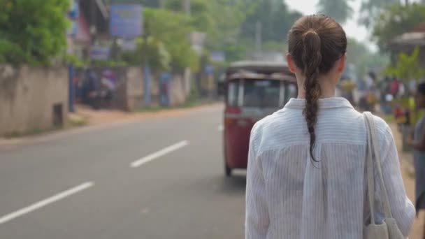 girl walks along road past moving vehicles in tropical city - Materiał filmowy, wideo