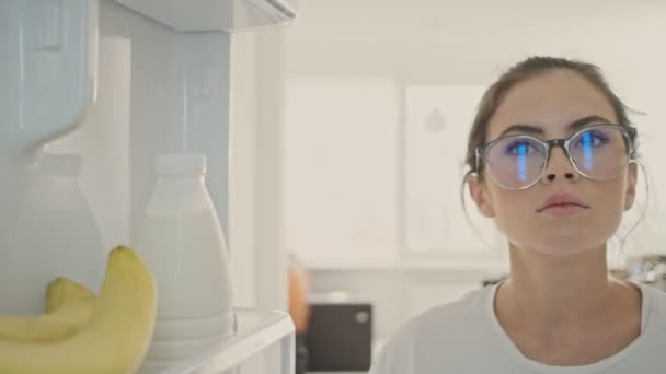 Calm pretty brunette woman in eyeglasses taking bottle of milk and bananas from refrigerator on kitchen - Materiał filmowy, wideo
