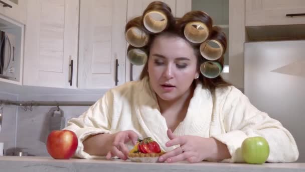 Plus-size girl, dressed in a bathrobe, curlers on her head, looks at the cake with desire, not daring to start eating it - Πλάνα, βίντεο