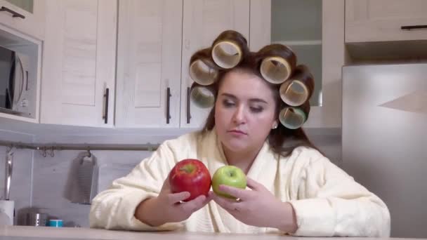 Plus-size girl, dressed in a bathrobe, curlers on her head, looks at apples, she does not want to eat them - Felvétel, videó