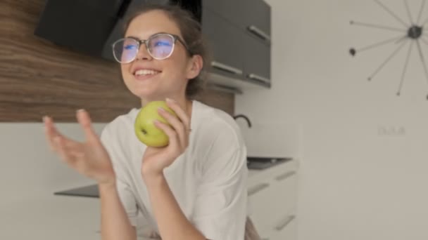 Весела красива брюнетка в окулярах грає з яблуком і дивиться навколо на кухню
 - Кадри, відео