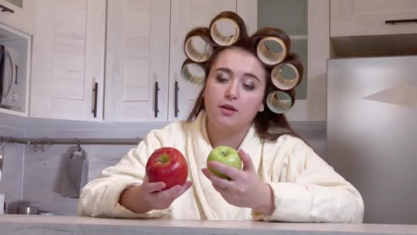 Plus-size girl, dressed in a bathrobe, curlers on her head, looks at apples, she does not want to eat them - Footage, Video