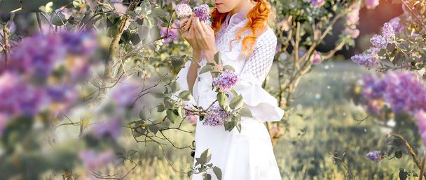 Beyaz elbiseli güzel kızıl saçlı beyaz kız bahar parkında leylak çiçeklerinin tadını çıkarıyor. - Fotoğraf, Görsel