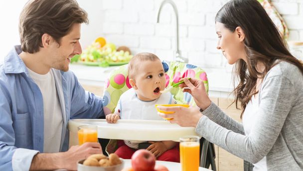 Jovem mãe e pai alimentando o bebê filho na cozinha
 - Foto, Imagem