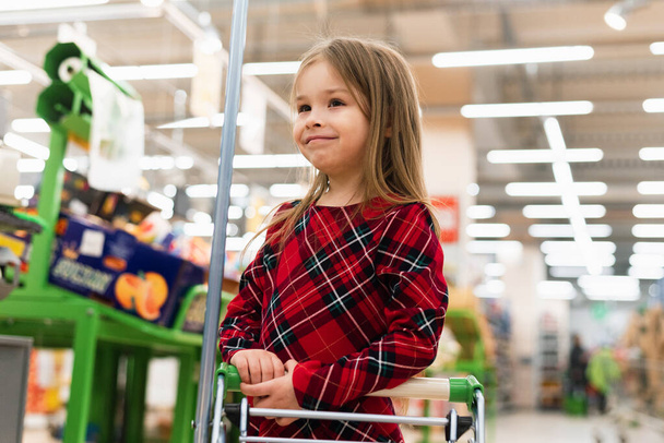 Ein fröhliches Mädchen hält einen Einkaufswagen in der Hand und wählt Produkte aus. Das Konzept des Einkaufs im Supermarkt, der Auswahl der Produkte, des Verkaufs. - Foto, Bild