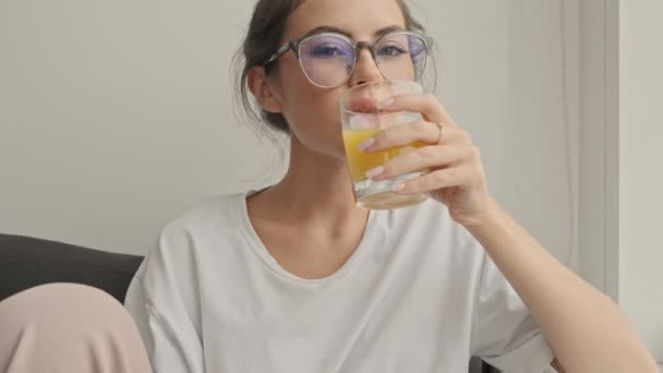 Boldog csinos barna nő szemüveg ivás gyümölcslé és a kamerába, miközben ül a kanapén otthon - Felvétel, videó