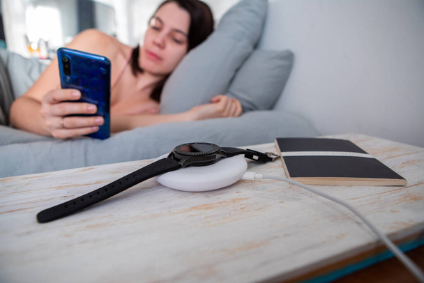 woman surfing internet on cellphone laying in bed smart watch on wireless charger copy space - Photo, Image