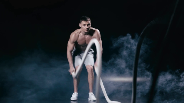 γυμνόστηθος άνθρωπος γυμνάζεται με σχοινιά μάχης σε μαύρο χρώμα με καπνό  - Πλάνα, βίντεο