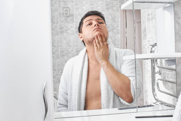 Ένας άντρας με λευκό μπουρνούζι κοιτιέται στον καθρέφτη στο μπάνιο.. - Φωτογραφία, εικόνα