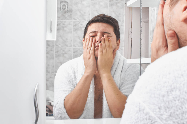 a man in a white bathrobe washes his face with soap through a bathroom mirror. - Foto, Imagen