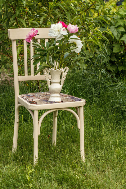 Witte rustieke stijl stoel en vaas met pioenachtige bloemen op groen gras in de tuin. - Foto, afbeelding