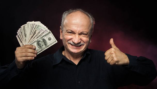 Caballero mayor sosteniendo una pila de dinero. Retrato de un anciano emocionado. Feliz anciano sosteniendo billetes de dólar sobre fondo rojo oscuro
.  - Foto, Imagen