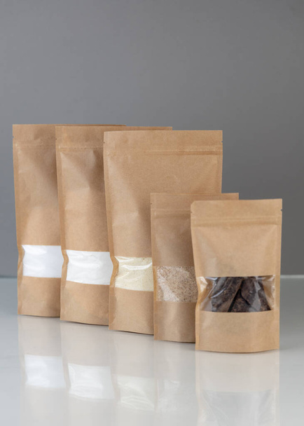 Barna kraft papír doypack táskák élelmiszerekkel szürke alapon. Csomagolás élelmiszerek és áruk mintadokumentumához. Tömegtermékekhez való ablakkal ellátott csomagolás. - Fotó, kép