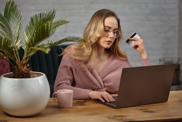 Female in pink cardigan, glasses. Working on laptop and showing white plastic card. Sitting at wooden table with pink cup, palm in pot on it. Close up - Foto, Imagem