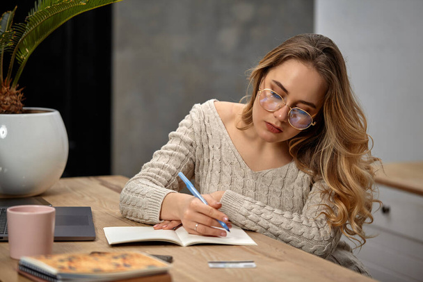 Blogueur modèle blonde en lunettes, pull beige. Assis dans la cuisine à la table en bois, va écrire quelque chose dans son carnet. Gros plan
 - Photo, image