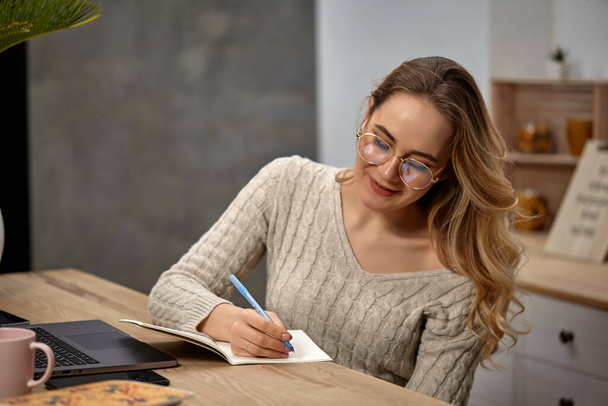 Blonde girl blogger in glasses, beige sweater. Smiling, sitting in kitchen at wooden table, going to write something in her notebook. Close up - Фото, изображение