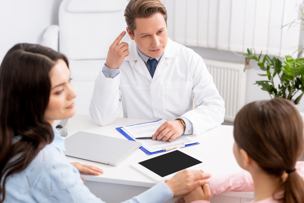 ent physician pointing with finger at ear near mother and daughter sitting at desk - Foto, imagen
