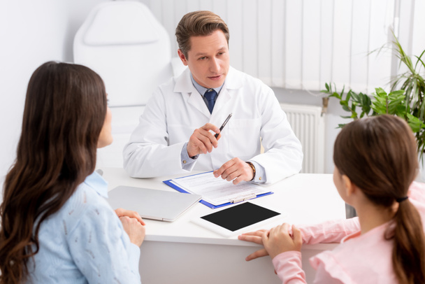 serious ent physician talking to mother and daughter near digital tablet with blank screen on desk - Foto, Imagen