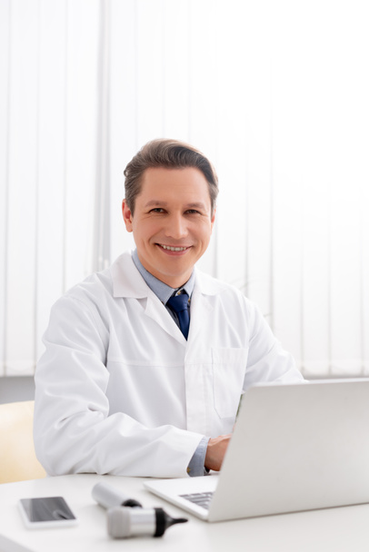 smiling otolaryngologist looking at camera while using laptop at workplace - Fotó, kép
