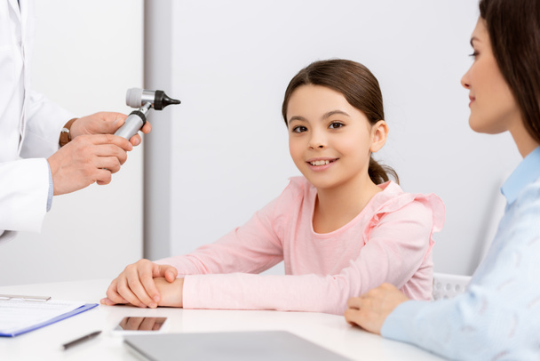 cropped view of otolaryngologist holding otoscope near smiling child and mother - Photo, Image
