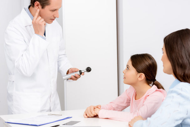 mother and daughter looking at otolaryngologist holding otoscope and pointing with finger at his ear - 写真・画像