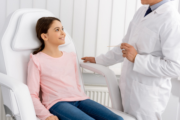 rajattu näkymä otolaryngologist tilalla kurkun lastalla seistessään lähellä hymyilevä lapsi istuu lääketieteellisessä tuolissa
 - Valokuva, kuva