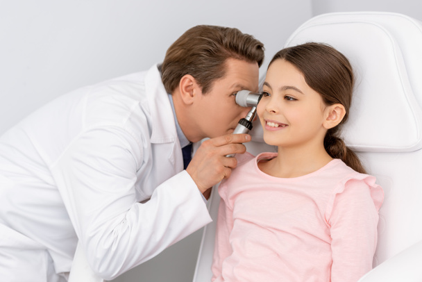 внимательный врач осматривает ухо улыбающегося ребенка с помощью отоскопа
 - Фото, изображение