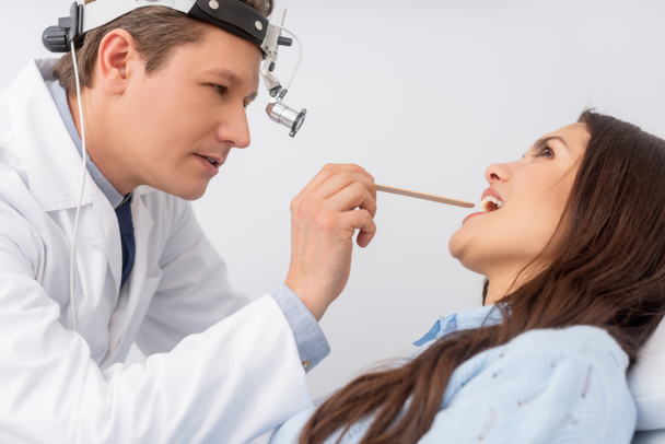 Aufmerksamer Hals-Nasen-Ohren-Arzt im Scheinwerferlicht, der den Hals eines attraktiven Patienten mit Kehlkopfspachtel untersucht - Foto, Bild