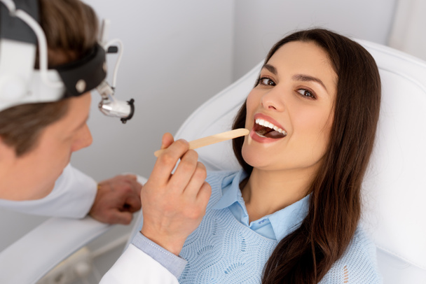 atrakcyjna kobieta patrząc w aparat podczas ent lekarz trzymając język szpatułka  - Zdjęcie, obraz