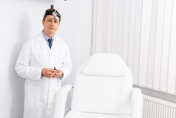 przystojny, pewny siebie otolaryngolog z reflektorem stojącym przy fotelu medycznym i patrzącym w kamerę - Zdjęcie, obraz