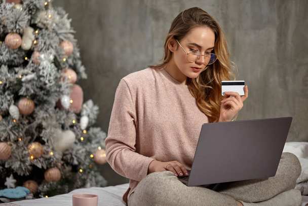 Blonde girl blogger in glasses, casual clothing. She is holding plastic card and working on her laptop, sitting on bed with cup. Close up, copy space - Photo, Image