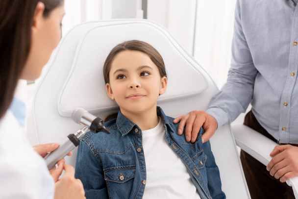 oříznutý pohled otce dotýkajícího se ramene dcery sedící v lékařském křesle poblíž otolaryngologa s otoskopem - Fotografie, Obrázek