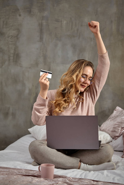 Blogger mulher loira em óculos, roupas casuais. Sorrindo, levantou o punho, segurando cartão de plástico branco, sentada na cama com laptop nas pernas. Close-up
 - Foto, Imagem