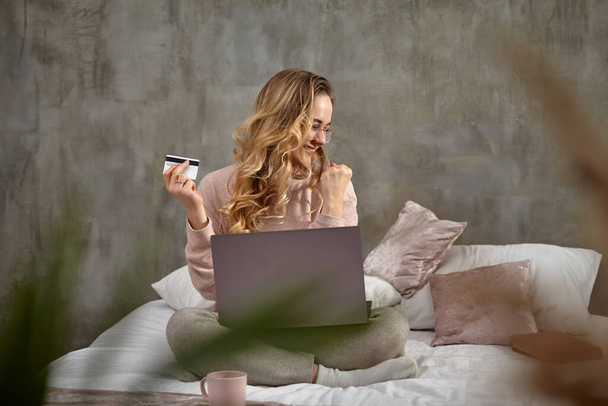 Blogger modelo rubia en gafas, ropa casual. Sonriendo, mirando emocionado, sosteniendo la tarjeta de plástico blanco, sentado en la cama con el ordenador portátil en las piernas. Primer plano
 - Foto, imagen