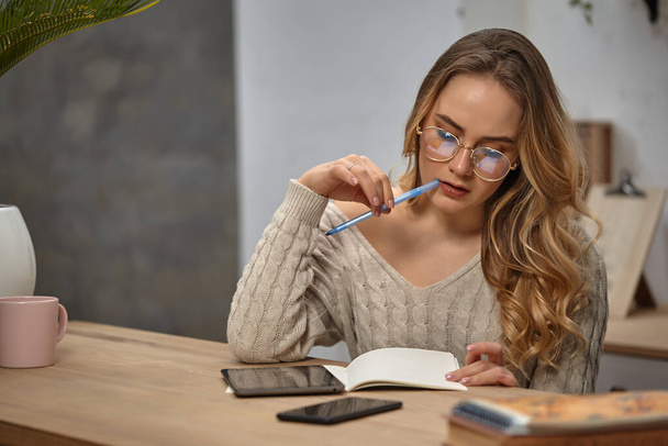 Vrouwelijke blogger met bril, beige trui. Zittend in de keuken aan tafel met beker, handpalm, tablet, smartphone en boeken. Ik ga iets schrijven. Close-up - Foto, afbeelding