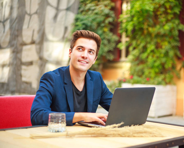 Charming young business man outdoors at a table in a summer cafe courtyard working at a laptop - Photo, Image